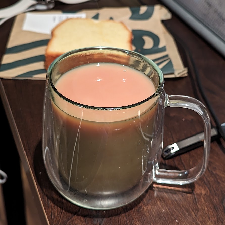 green rimmed glass mug of tea, sitting on desk, with a blurred lemon loaf visible behind the tea, square aspect ratio