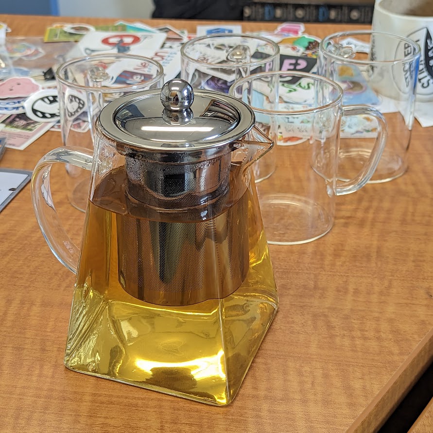 glass teapot, with infuser, currently steeping white tea, therefore the water is a warm yellow glow, behind kettle is three glass mugs, along with many different unapplied sticikers in the back of them