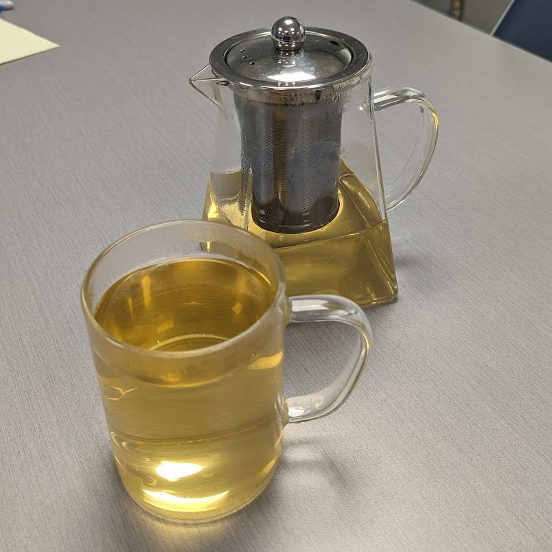 glass teapot, with infuser, containing steeped white tea, therefore the water is a warm yellow glow, infront of the kettle is a singular glass mug, which white tea has been poured into